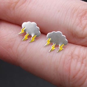 May include: A pair of silver-colored cloud earrings with yellow lightning bolt accents. The earrings are small and detailed, featuring a whimsical design. The image is a close-up, showcasing the earrings' design and craftsmanship.