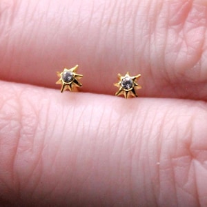 May include: A pair of small, gold stud earrings. Each earring has a starburst design with a central, clear gemstone. The earrings are shown on a person's finger against a dark background.