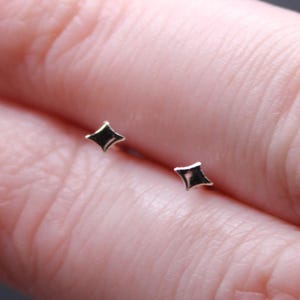 May include: Two tiny star-shaped stud earrings. Each earring has a black centre set within a gold-coloured frame. The earrings are shown on a person's finger, against a dark background.