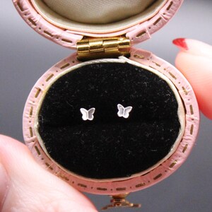May include: A small, open, pink jewellery box with gold hardware displays two tiny butterfly earrings on a black velvet interior. The box is held open, revealing the earrings. The background is a dark grey.