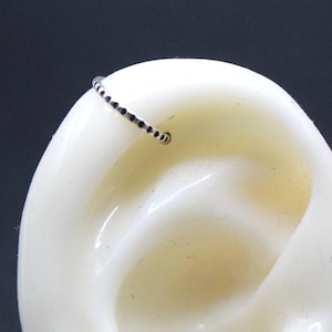 May include: A small, delicate silver hoop earring with black stones is placed on a white ear model. The earring is a simple, minimalist design. The ear model is held by a person with red nail polish.