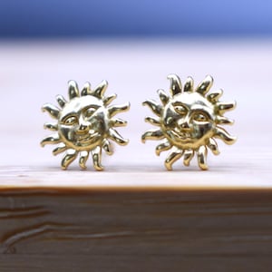 Könnte beinhalten: Ein Paar goldfarbene Sonnenohrringe mit einem Gesichtsdesign. Die Ohrringe liegen auf einer hellbraunen Holzoberfläche.