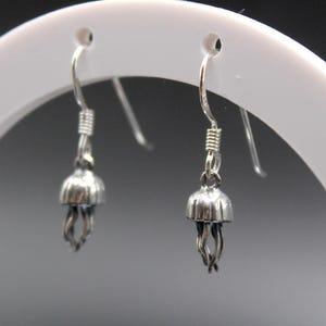 May include: A pair of silver-colored jellyfish earrings. Each earring features a small, bell-shaped top with delicate, dangling tentacles. The earrings are displayed on a white, circular stand against a dark background.