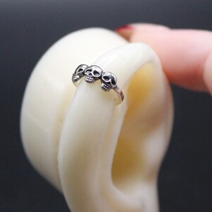 May include: A silver ring featuring three small skull designs. The ring is displayed on a white, curved object against a dark background. The ring is a piece of jewelry.
