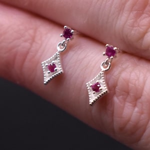 May include: A pair of silver earrings with a small red gemstone in the center of a diamond-shaped pendant.
