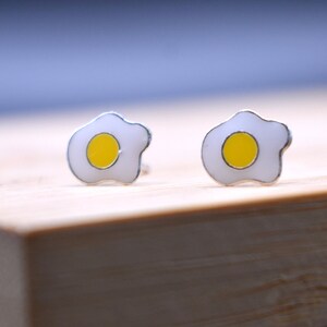 May include: A pair of silver earrings shaped like fried eggs with yellow yolks.