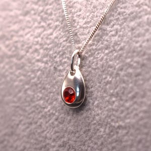 May include: A silver necklace featuring a teardrop-shaped pendant with a single, small, round, faceted red gemstone set in the centre. The pendant hangs from a thin, delicate silver chain that connects to a small silver jump ring at the top.