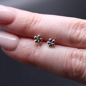 May include: Two small, silver-colored flower-shaped stud earrings. The earrings are held between fingers against a dark background. The earrings have a detailed, textured design, and are likely made of metal.