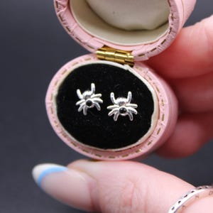 Könnte beinhalten: Ein Paar silberne Spinnen-Ohrstecker, präsentiert in einer rosa Schmuckschatulle mit schwarzem Samtinnenraum. Die Ohrringe haben ein detailliertes Spinnendesign mit kleinen schwarzen Akzenten. Die Schmuckschatulle ist geöffnet und zeigt die Ohrringe.