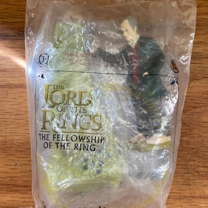 May include: A plastic bag containing a toy figure of a man wearing a dark coat and a red shirt. The bag has a Burger King logo and the text "The Lord of the Rings: The Fellowship of the Ring" printed on it.