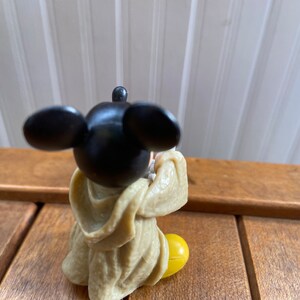 Puede incluir: Una peque&ntilde;a figura de pl&aacute;stico de Mickey Mouse con una t&uacute;nica marr&oacute;n y zapatos amarillos. La figura est&aacute; sentada con las manos juntas frente a ella.