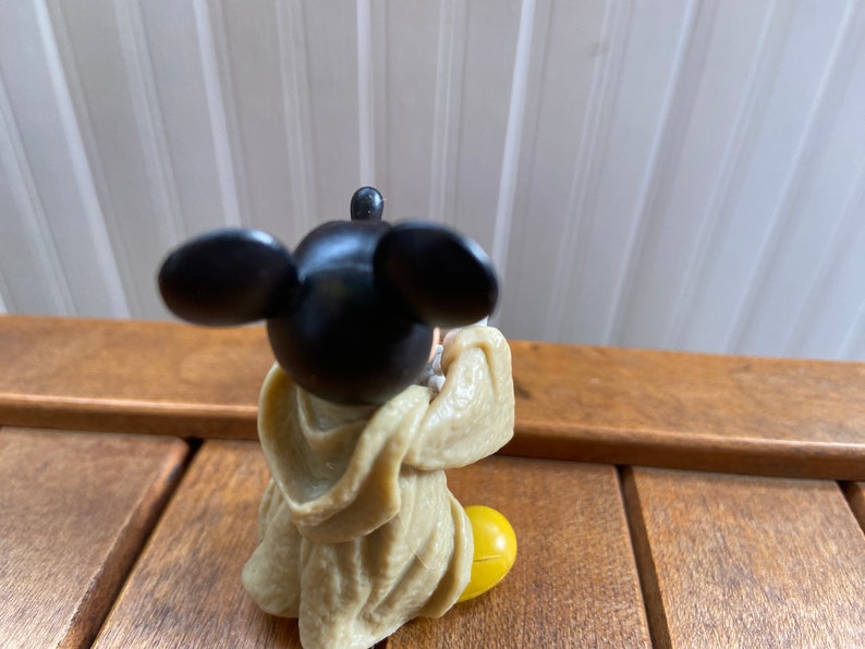 Puede incluir: Una peque&ntilde;a figura de pl&aacute;stico de Mickey Mouse con una t&uacute;nica marr&oacute;n y zapatos amarillos. La figura est&aacute; sentada con las manos juntas frente a ella.