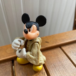 Puede incluir: Una figurita de pl&aacute;stico de Mickey Mouse con una t&uacute;nica de color beige. Mickey tiene una expresi&oacute;n seria en su rostro y est&aacute; sosteniendo sus manos levantadas frente a &eacute;l.