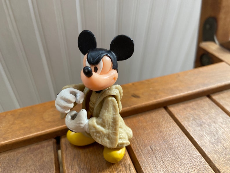 Puede incluir: Una figurita de pl&aacute;stico de Mickey Mouse con una t&uacute;nica de color beige. Mickey tiene una expresi&oacute;n seria en su rostro y est&aacute; sosteniendo sus manos levantadas frente a &eacute;l.