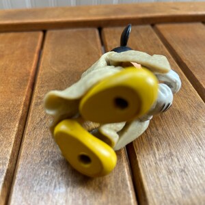 Puede incluir: Una figura de juguete de pl&aacute;stico amarilla y tostada de Mickey Mouse con un abrigo marr&oacute;n y zapatos negros. La figura est&aacute; tumbada de lado con los pies en el aire.