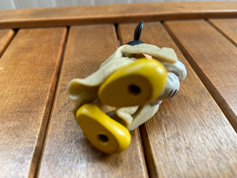 Puede incluir: Una figura de juguete de pl&aacute;stico amarilla y tostada de Mickey Mouse con un abrigo marr&oacute;n y zapatos negros. La figura est&aacute; tumbada de lado con los pies en el aire.