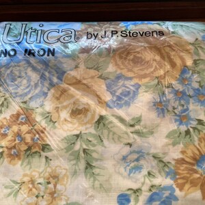 Puede incluir: Una sábana de tela blanca con un patrón floral en tonos azules, amarillos y marrones. La sábana está empaquetada y etiquetada "Utica by J.P. Stevens NO IRON".