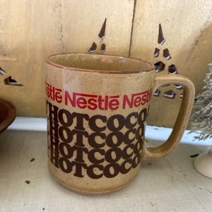 Peut inclure: Une tasse en céramique brune avec une anse. La tasse porte l'inscription "Nestle Nestle Hotcoco" en rouge et brun.