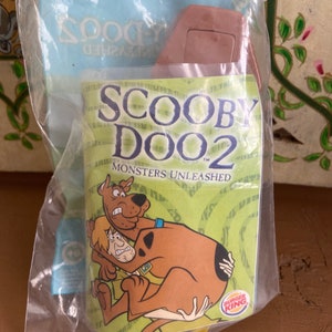 May include: A plastic bag containing a Scooby Doo 2: Monsters Unleashed toy. The toy is a brown plastic figure of Scooby Doo with a yellow collar. The bag also includes a Burger King logo.