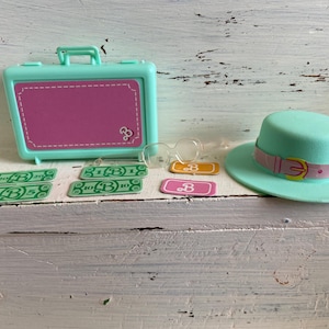 Puede incluir: Un conjunto de dinero de juego de Barbie, una maleta turquesa, un sombrero turquesa con una banda rosa y una hebilla, y un par de gafas transparentes. El dinero tiene la letra "B" y el número "10".