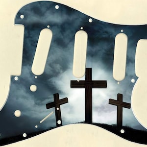 Puede incluir: Un pickguard de guitarra negro y blanco con un fondo de cielo tormentoso y tres cruces de madera en silueta contra las nubes.