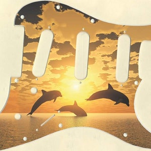May include: A guitar pickguard with a sunset and three dolphins jumping out of the water. The pickguard is made of a white material with a brown background.