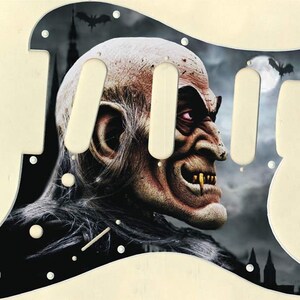 May include: Black and white guitar pickguard with a graphic of a monstrous creature with fangs and a red eye. The creature is set against a dark background with a full moon and bats.