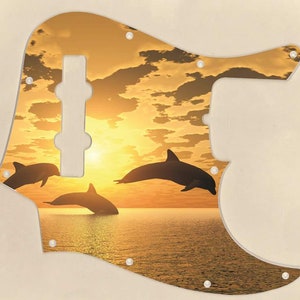 May include: A guitar pickguard with a sunset and dolphin silhouette design. The pickguard is a light brown color with a yellow sunset and three black dolphin silhouettes jumping out of the water.