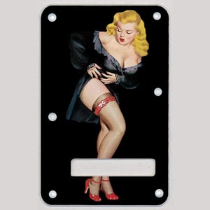 Könnte beinhalten: Ein schwarzer Gitarren-Pickguard mit einer Pin-up-Illustration einer Frau in einem schwarzen Kleid und roten Schuhen. Die Frau hält ihr Kleid hoch und hat blonde Haare. Der Pickguard hat ein leeres weißes Rechteck in der Mitte.