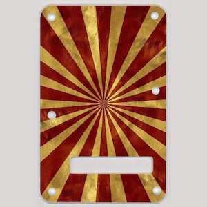May include: A red and gold guitar pickguard with a sunburst pattern. The pickguard is rectangular with rounded corners and four screw holes.