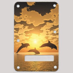 May include: Rectangular decorative panel featuring a sunset scene with three dolphins leaping out of the water. The sky is filled with golden clouds, and the sun is setting over the ocean. The panel has a rectangular cutout.