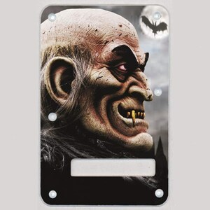 May include: A black guitar pickguard with a detailed illustration of a vampire with fangs and a menacing expression. The background is a dark, stormy sky with a full moon and a bat flying overhead.