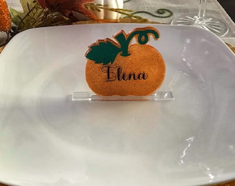 Puede incluir: Un plato cuadrado blanco con un borde dorado se encuentra sobre un plato de carga dorado. Un marcador de lugar con forma de calabaza con el nombre "Elena" está en el plato.