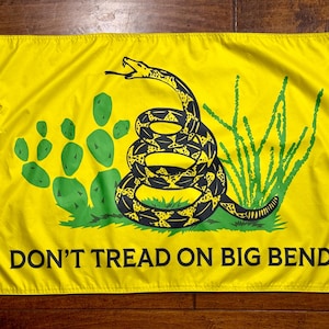 May include: A rectangular yellow flag featuring a coiled black and yellow rattlesnake in the center, flanked by a green prickly pear cactus and desert vegetation. The text DON'T TREAD ON BIG BEND is printed in black capital letters at the bottom.