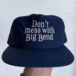 May include: A navy blue baseball cap featuring the embroidered white text "Don't mess with Big Bend" centered on the front panel. The cap has a structured crown, a flat brim with visible stitching, and a smooth, solid-colored fabric construction.