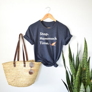 Könnte beinhalten: Ein dunkelgraues T-Shirt mit dem Text "Stop. Hammock Time." in weißen Buchstaben. Unter dem Text befindet sich eine kleine orangefarbene Illustration einer Katze in einer Hängematte. Das T-Shirt hängt an einem Holzbügel vor einer weißen Wand.
