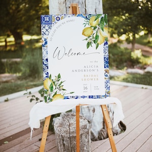 May include: A welcome sign for a bridal shower with a blue and white tile pattern and a lemon branch illustration. The text reads "So glad you're here!" and "Welcome to the Alicia Anderson's Bridal Shower" with the date "July 16, 20XX" and location "New York".