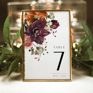 May include: A gold-framed table number card with a watercolor floral design in shades of burgundy, pink, and green. The card reads "Table 7" and "Bruna + Colin".