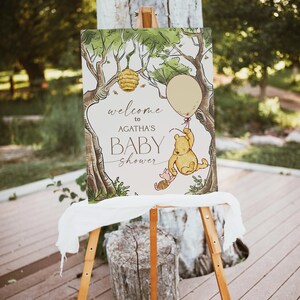 May include: A Winnie the Pooh themed baby shower welcome sign with a white background, green trees, a beehive, a honey pot, a balloon, Winnie the Pooh holding a balloon, and Piglet. The sign reads "Welcome to Agatha's Baby Shower".