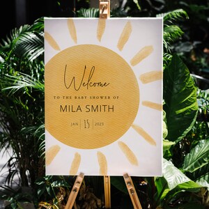 K&ouml;nnte beinhalten: Willkommen zur Babyparty von Mila Smith, ein wei&szlig;es Schild mit einer gelben Sonnengrafik und dem Datum 15. Januar 2023.