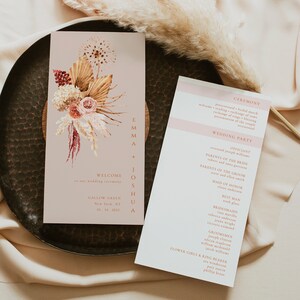 Pode incluir: Conjunto de convites de casamento em um esquema de cores rosa claro e creme. A frente do convite apresenta uma ilustração floral com flores secas e capim-dos-pampas. O verso do convite lista os detalhes da cerimônia e da recepção do casamento, incluindo o celebrante, os pais da noiva e do noivo, a dama de honra, o padrinho, as damas de honra e os padrinhos.