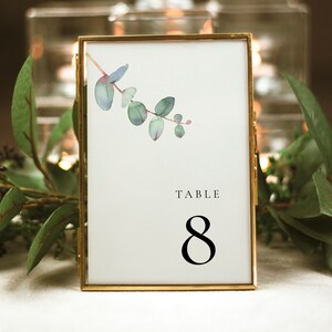 Può includere: Carta di numero tavolo con cornice dorata, un'illustrazione ad acquerello di foglie di eucalipto e il testo "TABLE 8".
