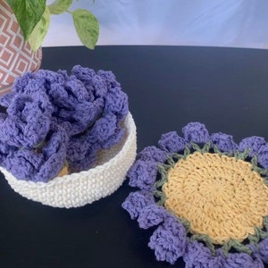 May include: A white crocheted basket filled with purple crocheted items, and a crocheted sunflower coaster with a yellow center and purple petals. The items are handmade and sit on a dark surface.