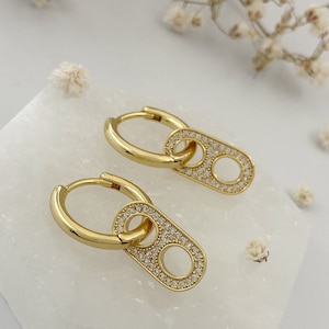 May include: A pair of gold hoop earrings with a dangling charm. The charm is a small oval shape with a smaller oval cut out in the centre. Both the hoop and the charm are covered in small clear rhinestones.
