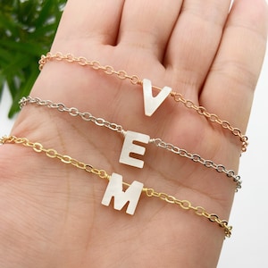 May include: Three delicate necklaces with white letter charms. The necklaces are layered on a hand, with the top necklace featuring a 'V', the middle necklace featuring an 'E', and the bottom necklace featuring an 'M'.