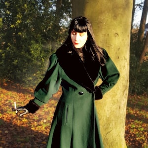 Puede incluir: Una mujer con un abrigo largo de color verde oscuro con cuello de piel negro. Está de pie en un bosque con hojas caídas en el suelo.