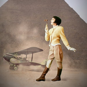 Op de afbeelding: Een vrouw in een geel jack en een bruine broek staat voor een grote piramide. Ze houdt een klein houten voorwerp in haar rechterhand. Een vintage-vliegtuig is geparkeerd in het zand op de voorgrond.