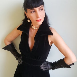 May include: A woman wearing a black velvet dress with a plunging neckline and long black gloves. She is wearing a pearl necklace and earrings.