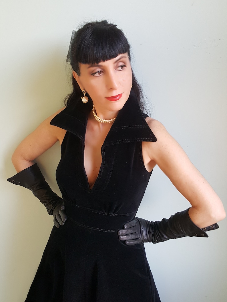 May include: A woman wearing a black velvet dress with a plunging neckline and long black gloves. She is wearing a pearl necklace and earrings.