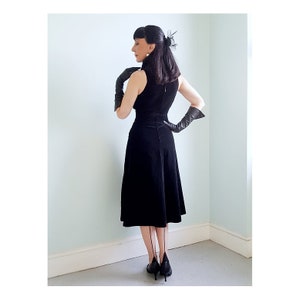 May include: A black velvet dress with a sleeveless bodice and a fitted waist. The dress has a flared skirt and a high neckline. The model is wearing black gloves and black heels.